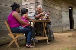 An older, white-haired Guatemalan women in brightly colored clothes uses a health chart to teach a younger mother with a child.