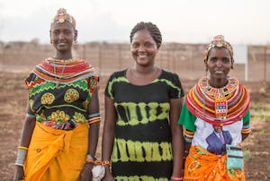 Three Kenyan women holding hands in traditional culture outfits and attire, with beaded necklaces and headbands