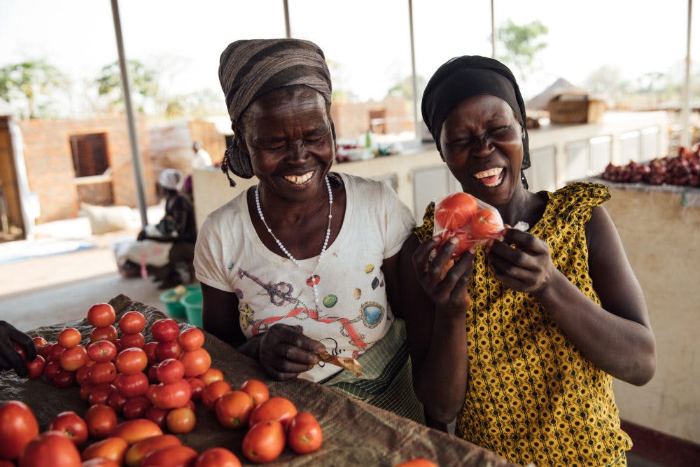 International Womens Day Photoessay - Two friends and women in Uganda smile in the local market next to ripe tomatoes