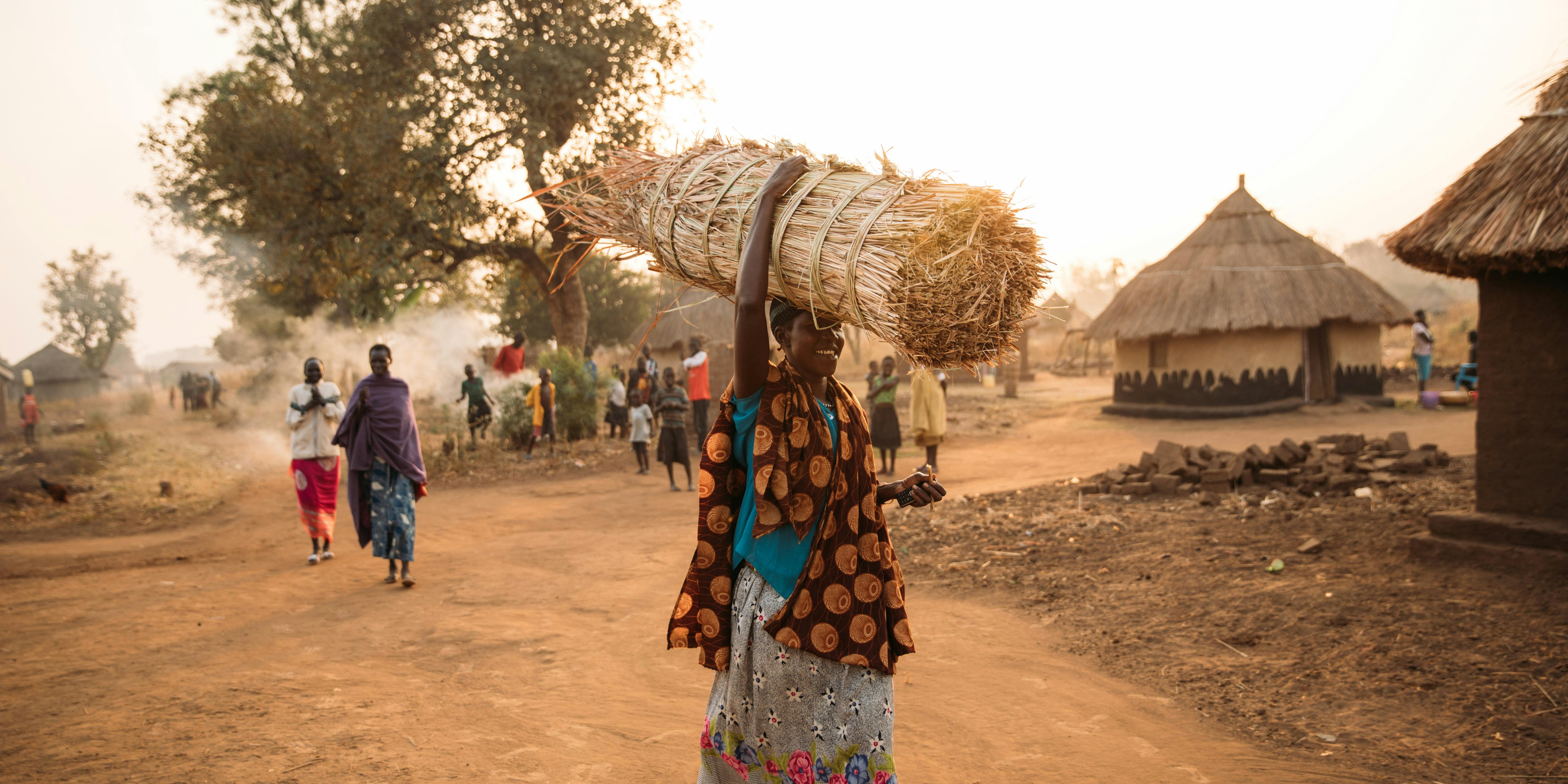 International Womens Day Photoessay - A woman in Uganda and flowy, traditional clothing carries a bundle of hay over her head as she walks across the dirt road in the village