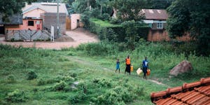 Landscape shot of a village in Uganda with three individuals carrying jerrycans down a road with lots of green grass.