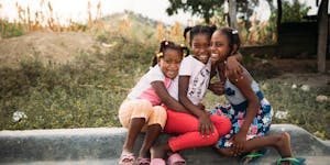Three girls in the Dominican Republic smiling and hugging, with mountain landscape in the background