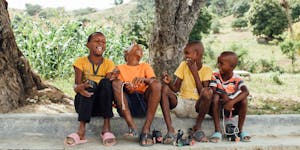 Boys in the Dominican Republic sit laughing on the curb, dressed in yellow and colorful shirts under a flourishing tree