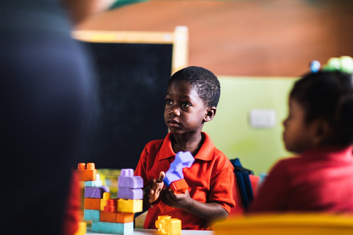 Dominican boy holds colorful building blocks that help with early childhood development, looking up at a teacher in a classroom