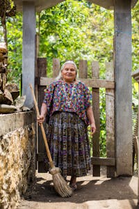 Elderly Guatemalan woman in traditional clothing stands outside her house with a broom