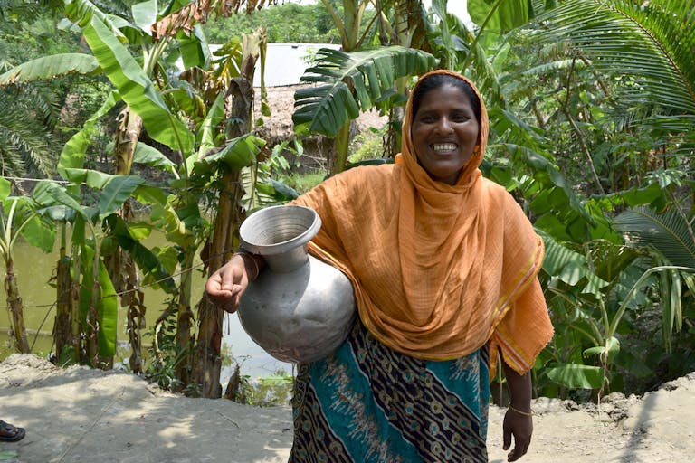 Woman in orange sari in Bangladesh holding a metal jug smiling