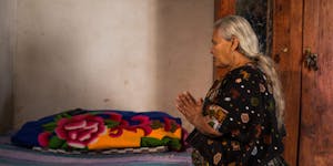 Elderly Guatemalan woman kneels at her bed and prays, with traditional embroidered blanket on the bed.
