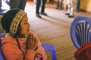 a child with a hat on is praying with her hands together and eyes closed
