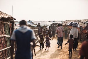 children and adults walking on dusty path between refugee homes