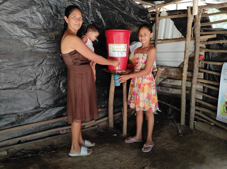Mother with baby and older daughter drawing water from bucket with filter
