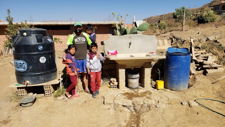 Man with two children standing between plastic water storage tank and cement sink
