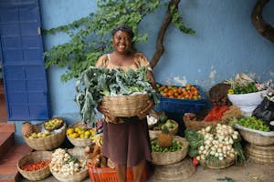 Woman wearing skirt and head scarf holding a large bushel of vegetables