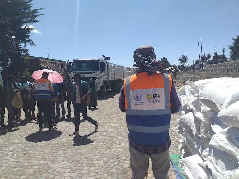 man with back to camera with Food for the Hungry and World Food Programme logos on his back, looking down a road with a delivery truck convoy parked