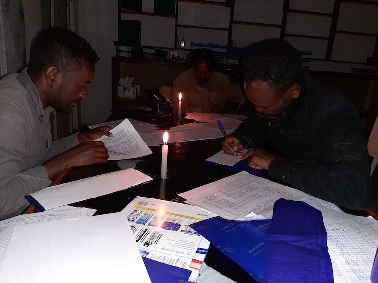 Men in a dark room with two candles on the table between them, working on paperwork