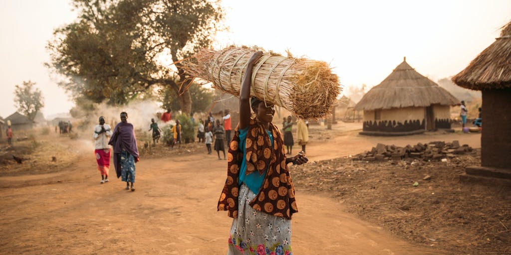 A woman in Uganda and flowy, traditional clothing carries a bundle of hay over her head as she walks across the dirt road in the village